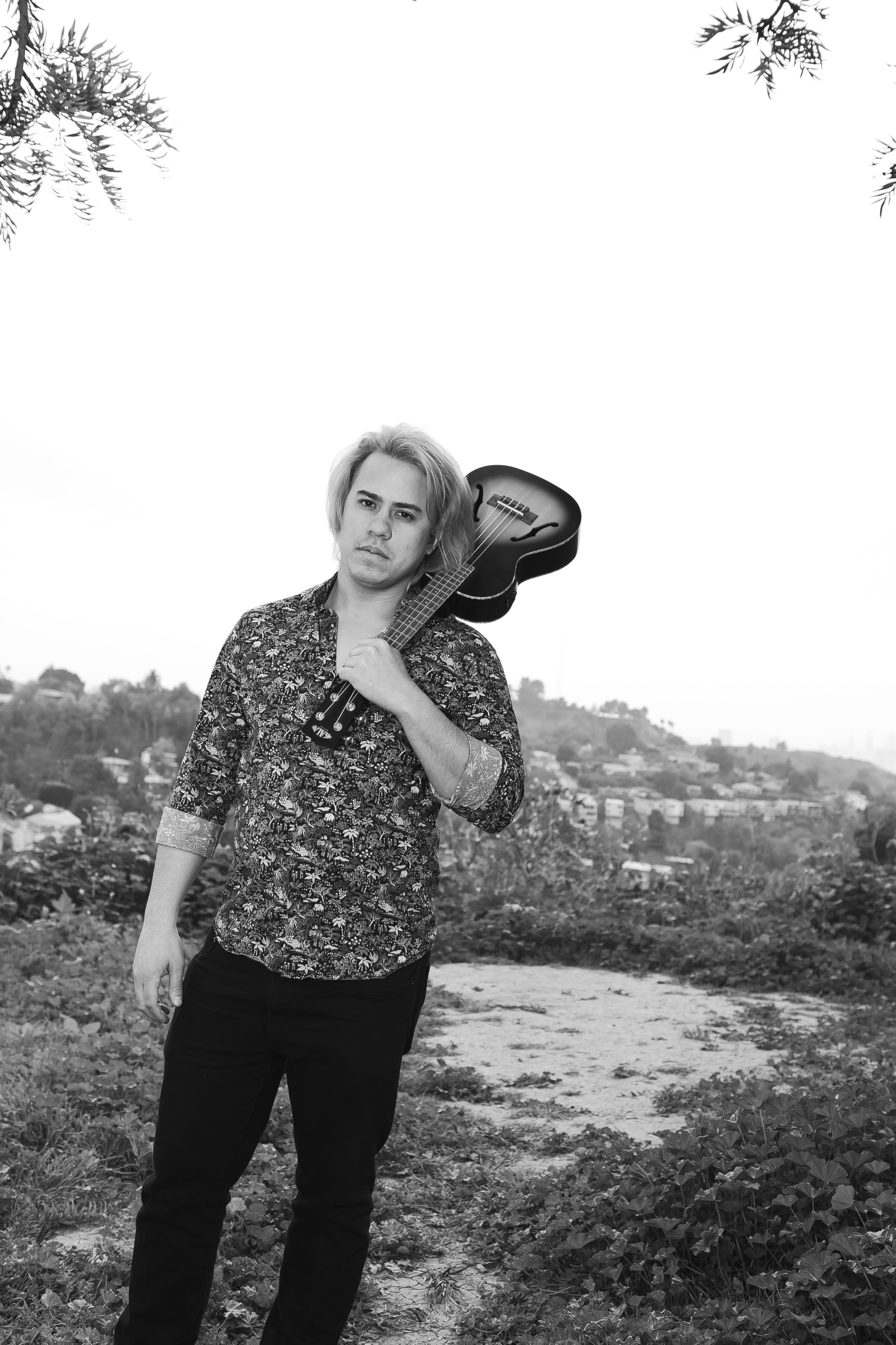 A man with blonde hair wearing a floral shirt stands outdoors, holding a guitar over his shoulder, with a blurred cityscape in the background.